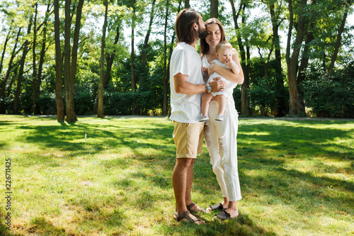 Dark-haired father kisses his beautiful wife holding a small daughter in a white dress in a park on a sunny day.