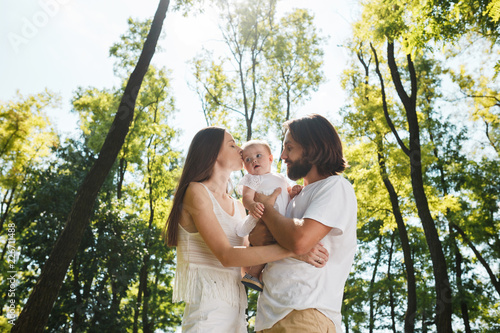 Happy family on the open air. Young dark-haired woman and her husband are kissing their charming little daughter. © Leika production