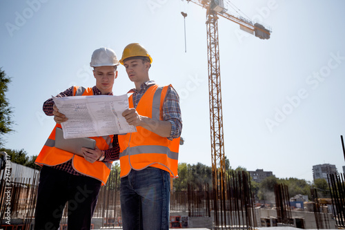 Architect and construction manager dressed in orange work vests and helmets discuss documentation on the open air building site next to the crane