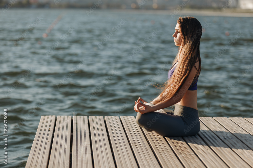 Young beautiful girl with long brown hair in a sports top and tights sits in lotus position on the wooden pier near the water on the summer day