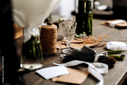 There are wineglass, little bouquet of flowers, postcard, envelopes, bobbin of twine and white ribbon on the wooden table