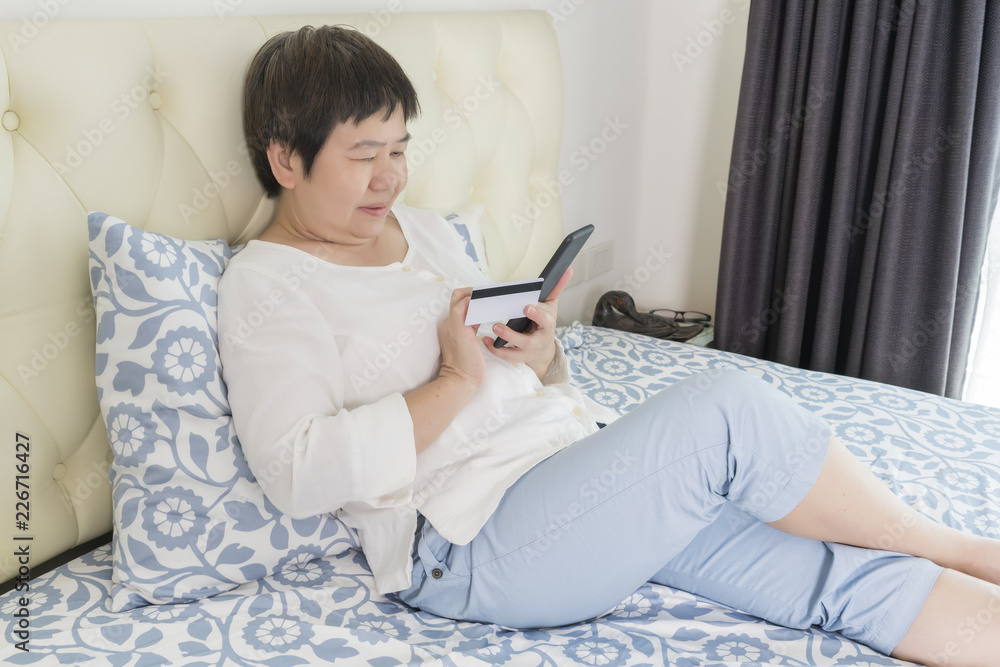 Asian middle-aged woman using smart phone and credit card for paying bills or shopping online in the bedroom Asian middle-aged woman using smart phone and credit card for paying bills or shopping online in the bedroom