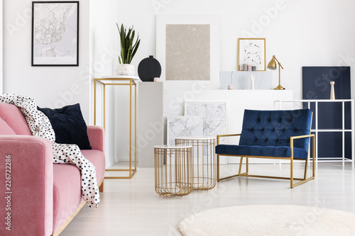 Silver abstract painting on the wall of trendy living room with two elegant coffee tables, petrol blue armchair and powder pink couch with doted blanket on it