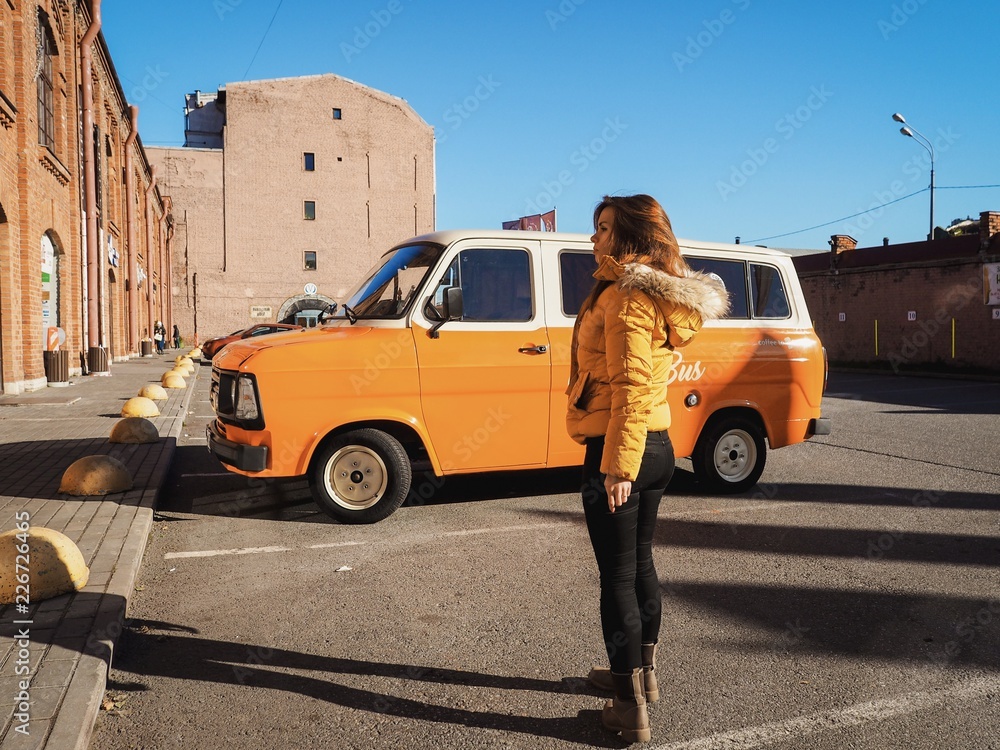 A girl with long hair in a bright yellow jacket stands near an orange mini bus in the middle of the street