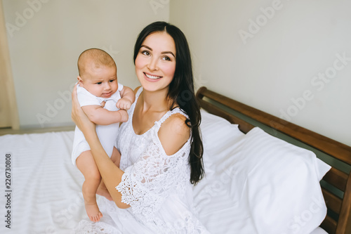 smiling young mother carrying her adorable liitle baby and sitting on bed at home