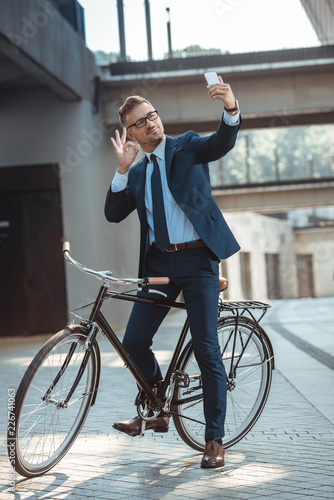 smiling businessman showing ok sign to smartphone while sitting on bicycle on street