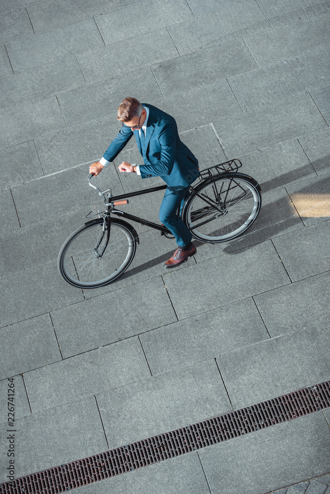 Obraz premium high angle view of businessman in formal wear riding bicycle and checking wristwatch