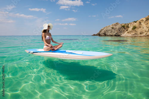 Young beautiful woman meditating in a sea at SUP paddleboarding. Healthy lifestyle. Girl in yoga pose relaxing in calm water.