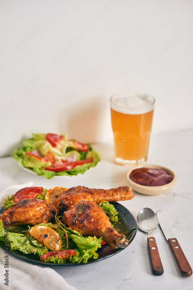 Grilled chicken legs roasted on the grill on dark plate with tomato sauce in a bowl and lettuce leaves, glass mug of beer