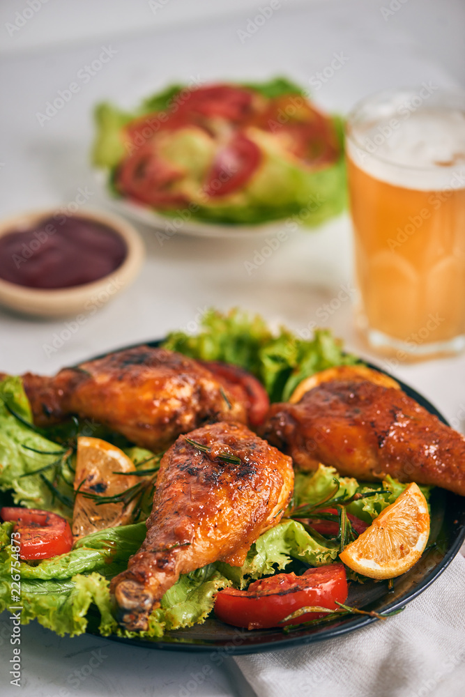 Grilled chicken legs roasted on the grill on dark plate with tomato sauce in a bowl and lettuce leaves, glass mug of beer Grilled chicken legs roasted on the grill on dark plate with tomato sauce in a bowl and lettuce leaves, glass mug of beer