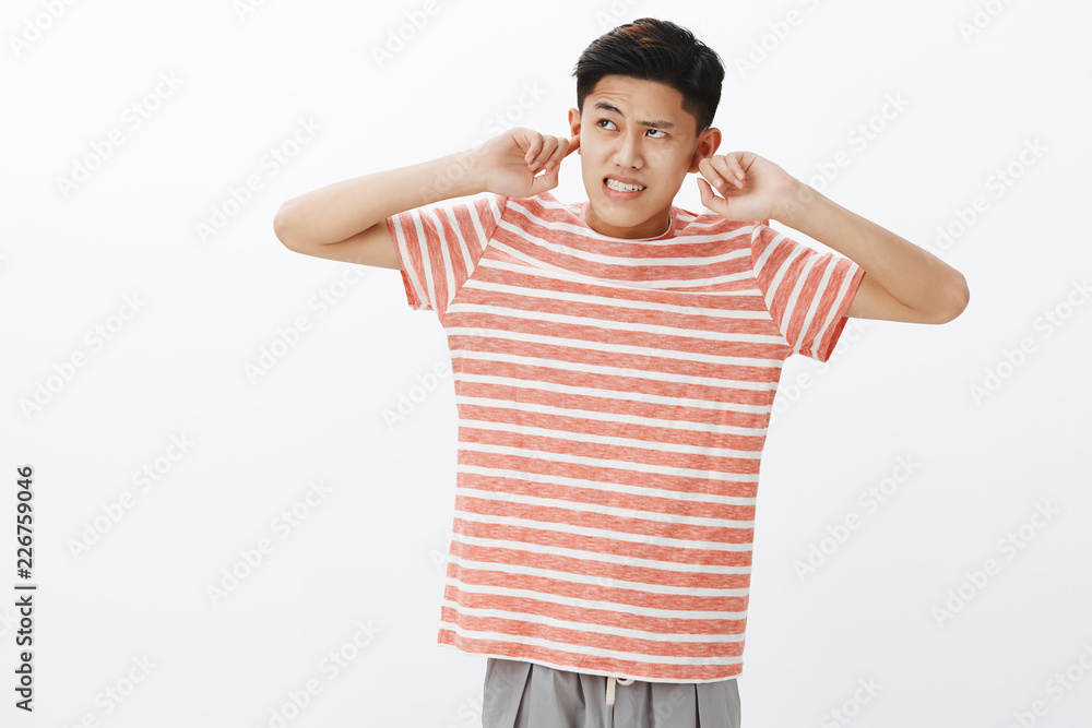 Young guy cannot focus on homework as hearing irritating and disturbing  loud noise coming from upstairs clenching teeth and frowning annoyed,  closing ears with index fingers, looking up angry Stock Photo