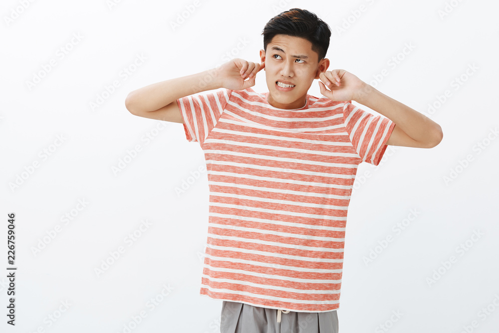 Fototapeta premium Young guy cannot focus on homework as hearing irritating and disturbing loud noise coming from upstairs clenching teeth and frowning annoyed, closing ears with index fingers, looking up angry