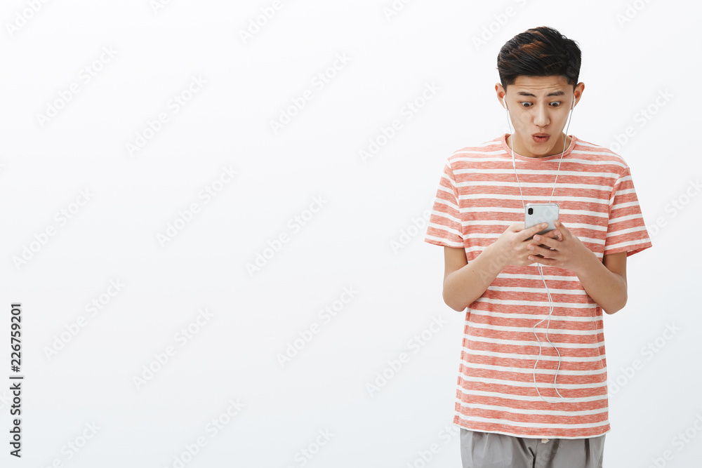 Impressed happy excited attractive young asian male with cool hairstyle in striped t-shirt holding smartphone looking pleased and amazed at cellphone screen saying wow, posing over white wall Impressed happy excited attractive young asian male with cool hairstyle in striped t-shirt holding smartphone looking pleased and amazed at cellphone screen saying wow, posing over white wall