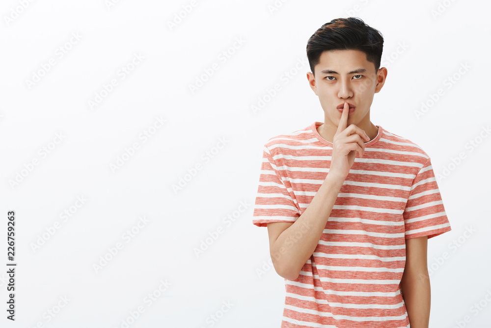 Asian elder brother looking serious and strict at camera demanding keep word and never tell anyone secret making shush gesture with index finger over closed lips, having mystery over white background Asian elder brother looking serious and strict at camera demanding keep word and never tell anyone secret making shush gesture with index finger over closed lips, having mystery over white background