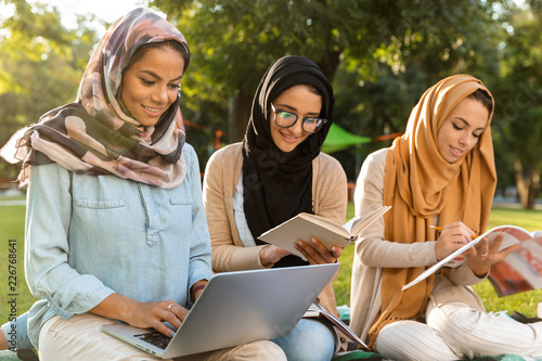 Canvas Print Women students using laptop computer and holding books in park.