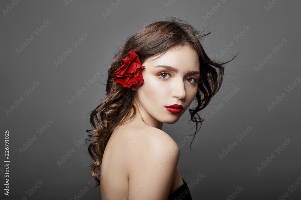 Fototapeta premium Woman with a big red flower in her hair. Brown-haired girl with a red flower posing on a gray background. Big beautiful eyes and natural makeup. Long curly hair, perfect face