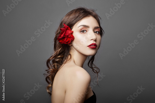 Woman with a big red flower in her hair. Brown-haired girl with a red flower posing on a gray background. Big beautiful eyes and natural makeup. Long curly hair  perfect face