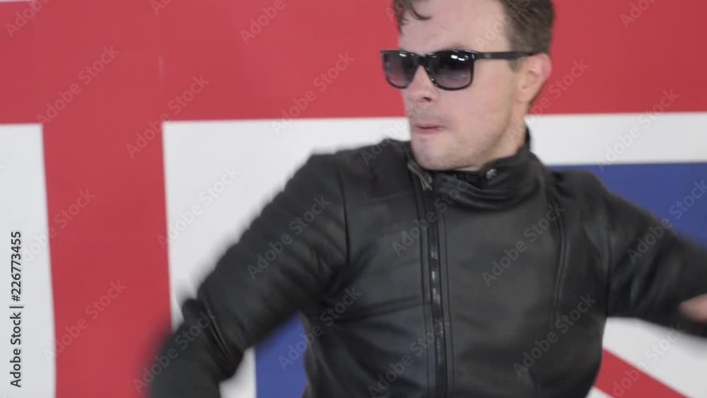 Cute charismatic caucasian dark-haired male wearing sunglasses and black leather motorcycle jacket with pants is actively dancing and singing on camera in studio with background of large UK flag. Cute charismatic caucasian dark-haired male wearing sunglasses and black leather motorcycle jacket with pants is actively dancing and singing on camera in studio with background of large UK flag.