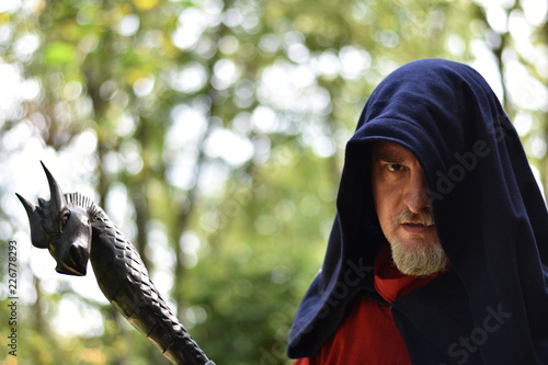 Close-up of a caucasian man over 40 wearing an hooded cloak and holding a black wooden  staff topped by a dragon head. The hood hides his left eye, could be a wizard or even  Odin in disguise.