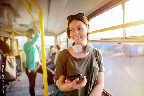 Modern smiling cute girl standing in a bus with headphones around her neck. Smiling and looking away.