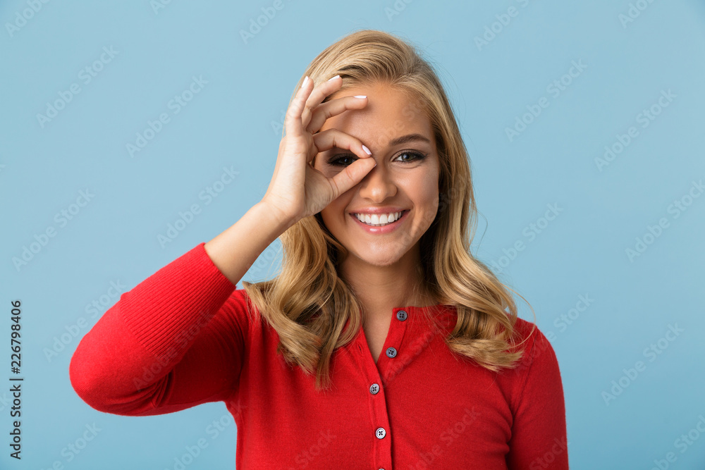 Portrait of cute blond woman 20s wearing red shirt smiling and looking through hole made by fingers, isolated over blue background in studio Portrait of cute blond woman 20s wearing red shirt smiling and looking through hole made by fingers, isolated over blue background in studio