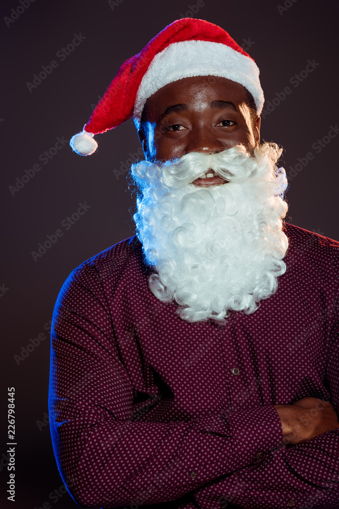 smiling african american man in santa hat and beard posing with crossed arms on black