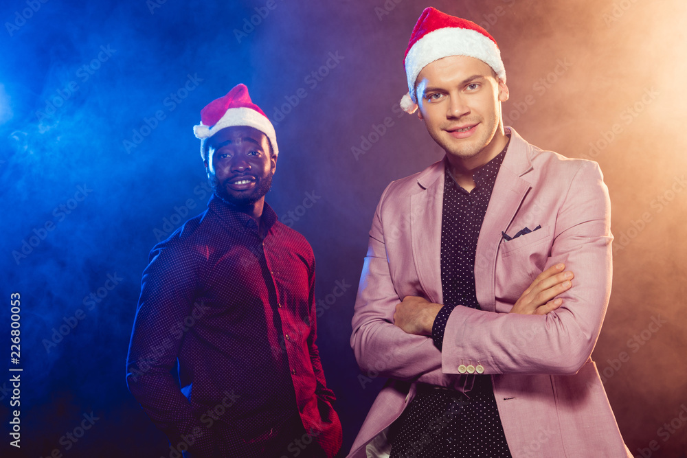 handsome man in pink jacket and santa hat standing near african american friend on black with smoke and backlit handsome man in pink jacket and santa hat standing near african american friend on black with smoke and backlit