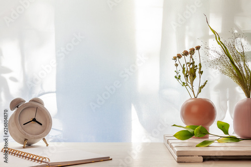 Photo of a desk with a concrete clock, an open notebook and vases with dried grasses on a background of sun-lit curtains