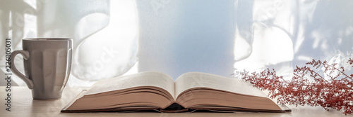 Panoramic photo of a desk with an open book, dried red grass and a gray cup with coffee on a background of sun-lit curtains