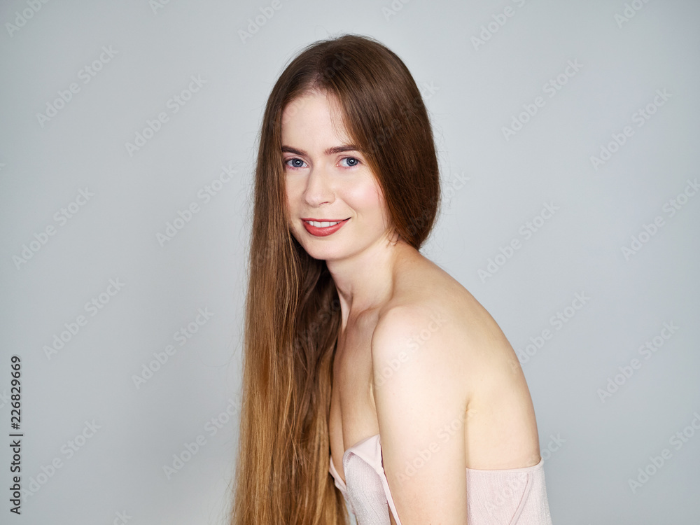 Natural light portrait of young beautiful blond woman with long hair in vintage dress smiling looking at camera over grey wall background.