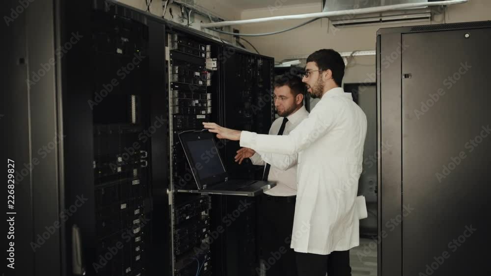 Two IT engineers are working in a data center with rows of server racks and super computers. They are discussing their work as they check cables and other equipment. Two IT engineers are working in a data center with rows of server racks and super computers. They are discussing their work as they check cables and other equipment.