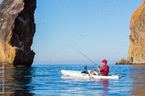 A man fishing on a kayak boat in the sea near the rocks at the shore of island mountain. Fisherman kayaking. Young fisherman kayaking.