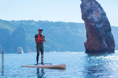 A man fishes on a fishing tackle in the standup paddleboard. Young fisherman on the SUP surfing in the sea near the island with rocks and mountains.