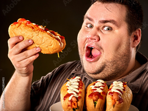 Hot dog contest. Fat man eating fast food hot dog on plate. Breakfast for overweight person. Junk meal leads to obesity. Dangers of extra weight. Person regularly overeats concept on black background.