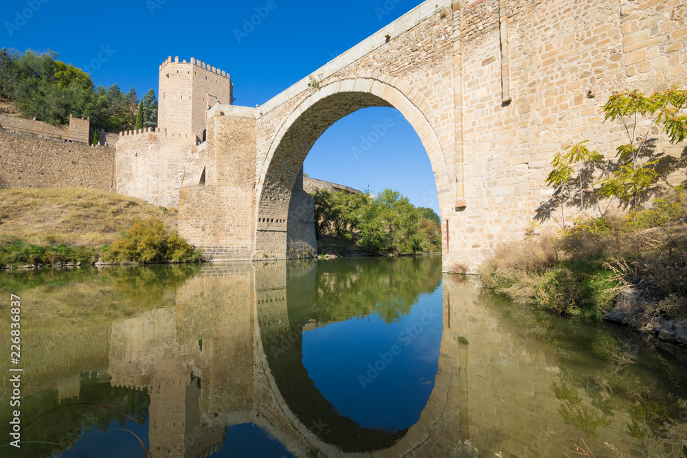 horizontal shot of arch of Alcantara bridge, landmark and monument from ancient Roman age, reflected on water of river Tagus, Tajo in Spanish, in Toledo city, Spain, Europe