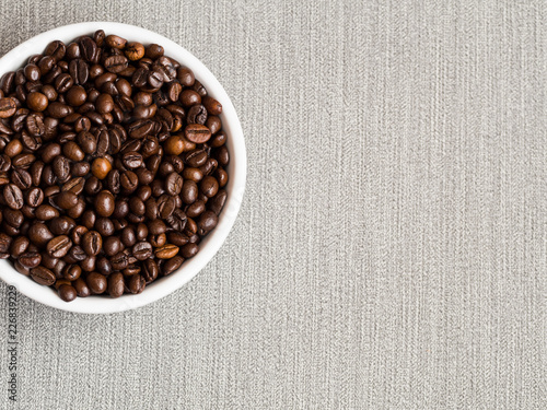 Coffee beans in white bowl on gray textile background. Top view, copy space, macro