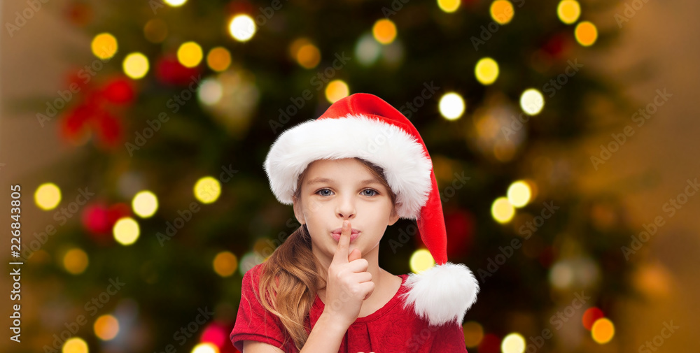 holidays and children concept - happy girl in santa hat making shh gesture over christmas tree lights background holidays and children concept - happy girl in santa hat making shh gesture over christmas tree lights background