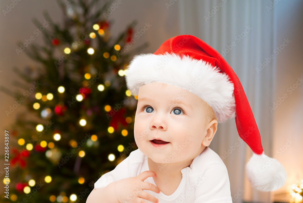 holidays, children and people concept - close up of happy little baby boy or girl in santa hat over christmas tree lights background