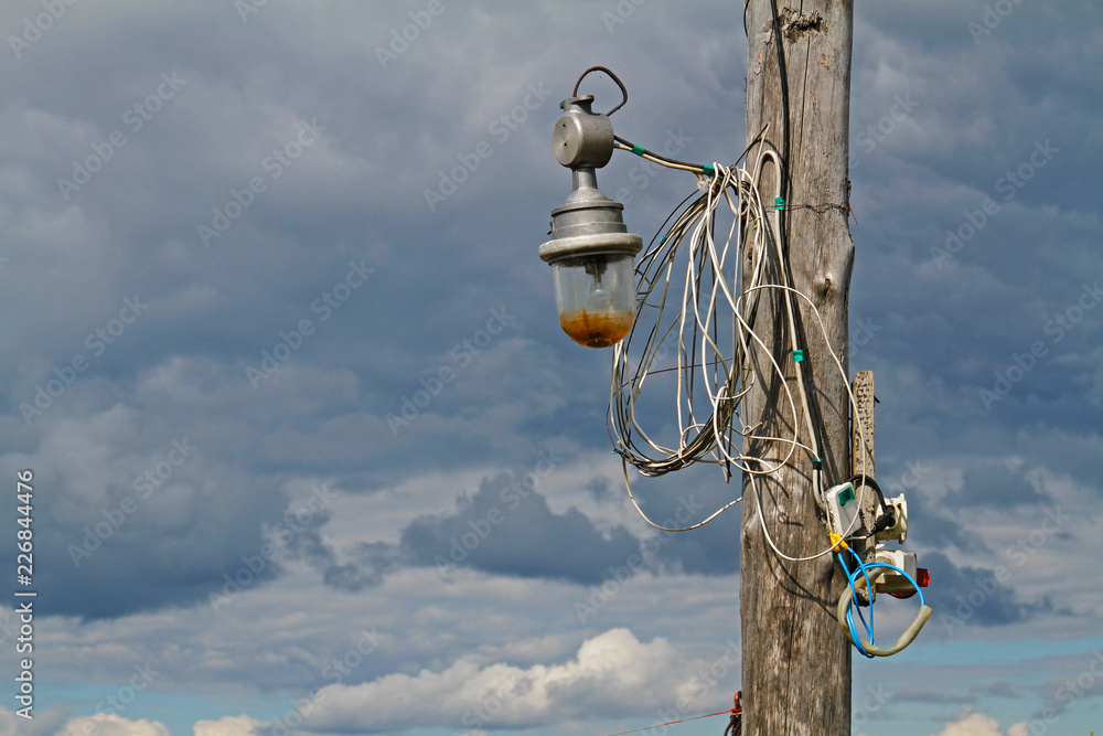 Fototapeta premium Wooden pillar with old lantern and wires against the sky with clouds
