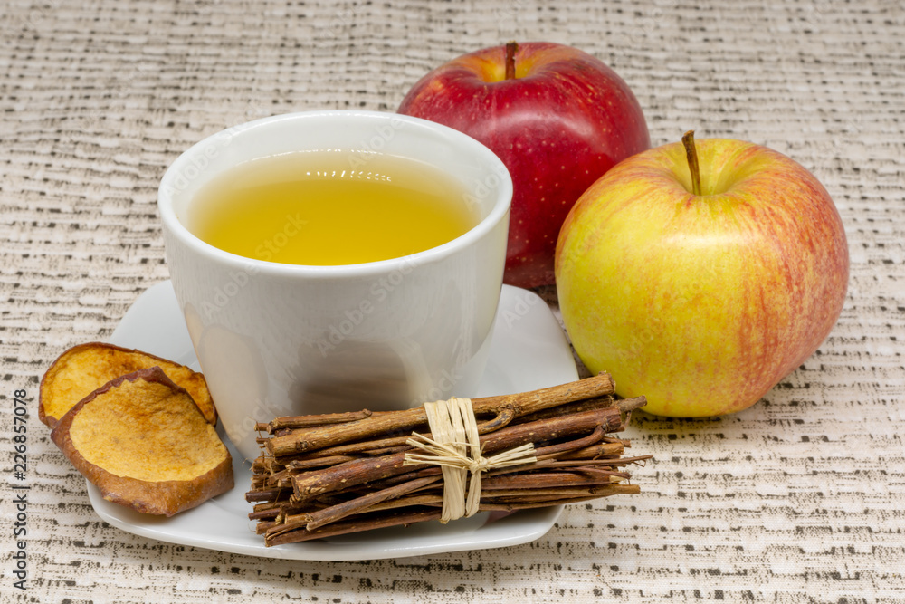 A cup of apple tea with two apples and dehydrated slices of the fruit with a patterned table cloth in the background and a bunch of cute little sticks A cup of apple tea with two apples and dehydrated slices of the fruit with a patterned table cloth in the background and a bunch of cute little sticks
