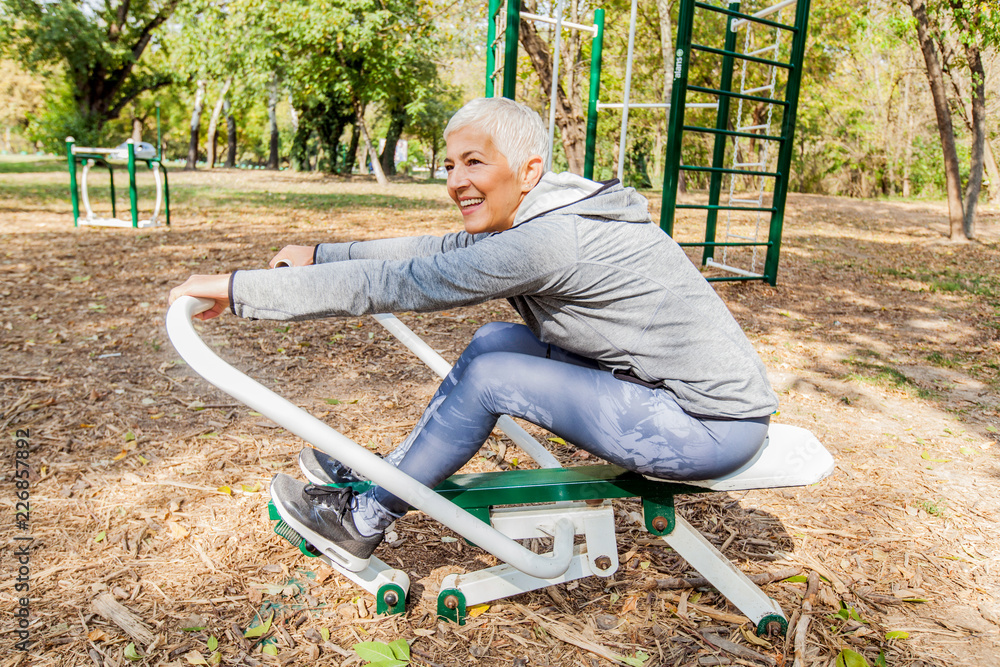 Elderly Woman Exercising At Outdoor Fitness Park With Rowing Machine Elderly Woman Exercising At Outdoor Fitness Park With Rowing Machine