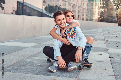 Trendy dressed young couple - pretty girl embrace her boyfriend while they sitting together on a skateboard near skyscraper. © Fxquadro