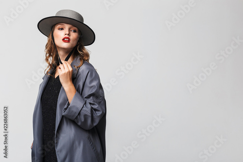 Young beautiful woman with wavy hair in modern hat and cloak thoughtfully looking aside over gray background isolated