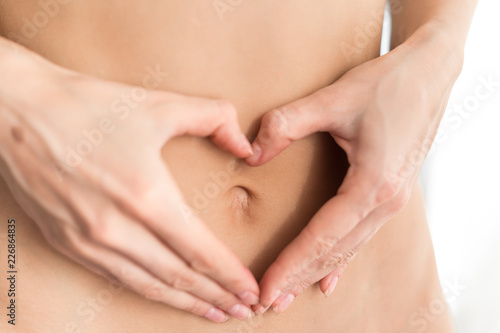 Caucasian woman without clothes holds fingers in the form of a figure a heart before a stomach. Light background