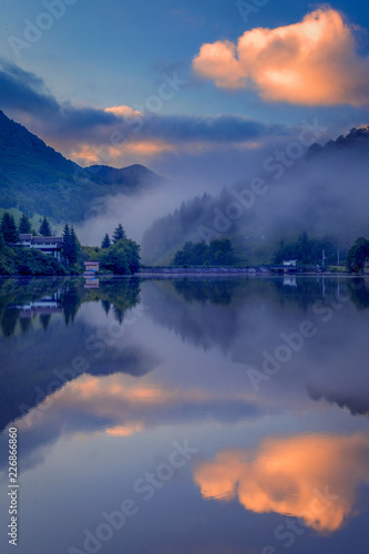 Foggy morning on a lake in the mountains with some houses on the shore with beautiful reflections in theb water