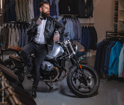 Stylish tattooed bearded man with dressed in black leather jacket and bow tie posing near retro sports motorbike at men's clothing store. © Fxquadro