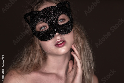 Beautiful young tender woman posing isolated over dark beige chocolate brown background wall wearing carnival cat masquerade mask looking camera.
