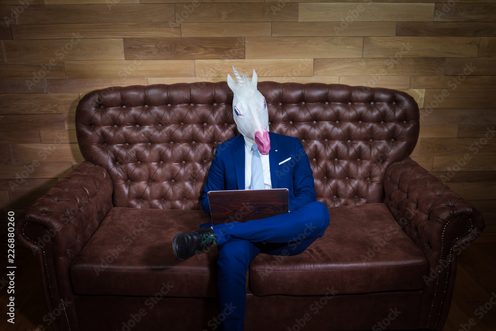 Strange man in elegant suit working at home office. Unusual young manager in comical mask on background of wooden wall. Funny unicorn sits on sofa like a boss. - obrazy, fototapety, plakaty 