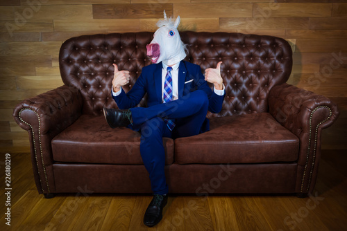 Funny unicorn in elegant suit sits on sofa like a boss and showing gesture thumbs up. Portrait of unusual man at home. Freaky guy in comical mask.