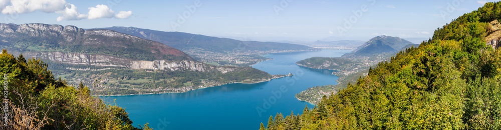 Panoramic view from Lake Annecy in Haute Savoie, France. The turquoise lake is lit by the sun. Some boats are travelling on the water.